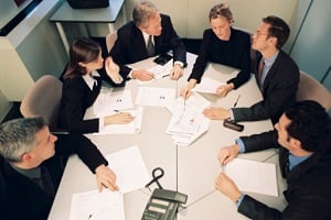 A group of individuals wearing suits sitting around a conference room table with papers in front of them.