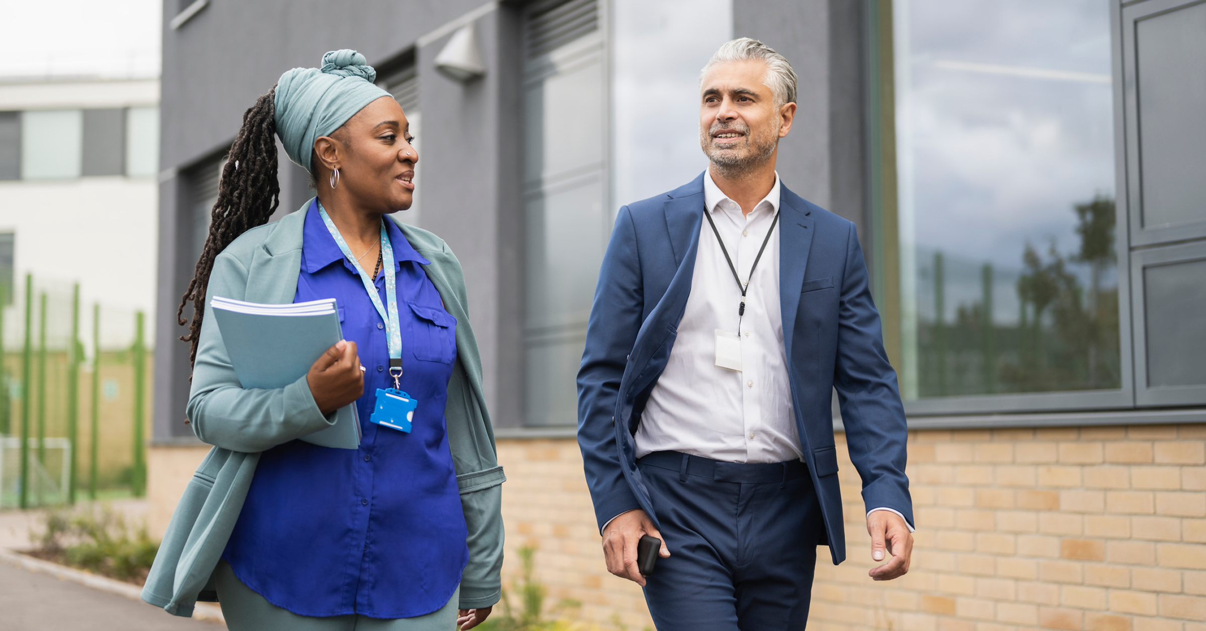 woman-in-blue-shirt-holding-tablet-walking-with-man-in-business-attire-outside-modern-building