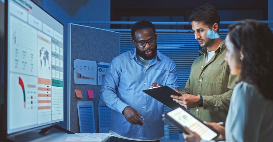 A team looks over a white screen and clipboard displaying busienss data. 