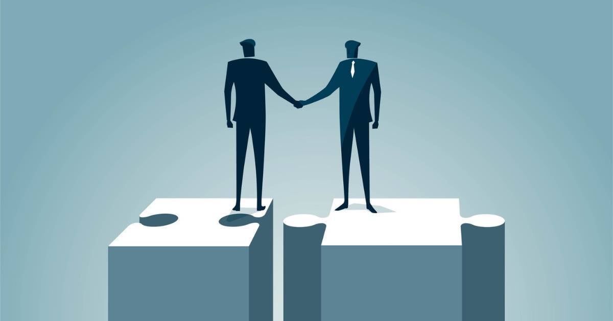 TWo business leaders stand on a puzzle piece shaking hands.