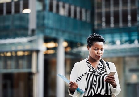 A business woman walking outside, looking at her cell phone and holding important documents. 