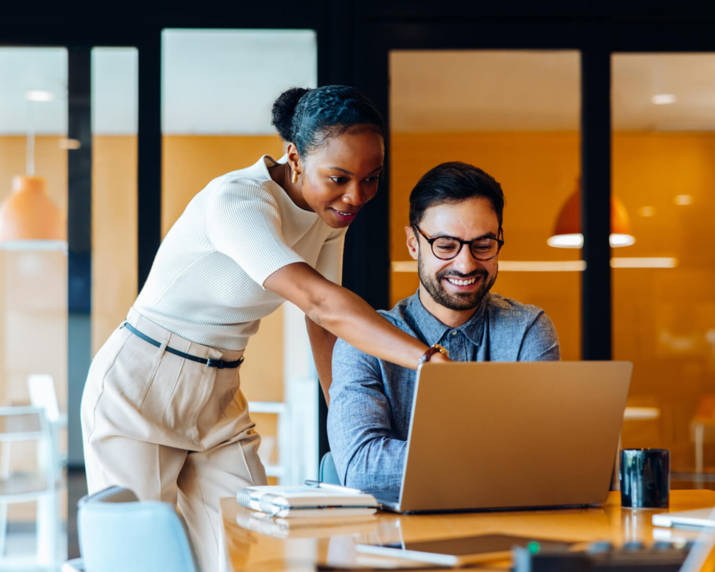 Two colleagues, a man and a woman, smiling while reviewing work together on a laptop in a modern office.