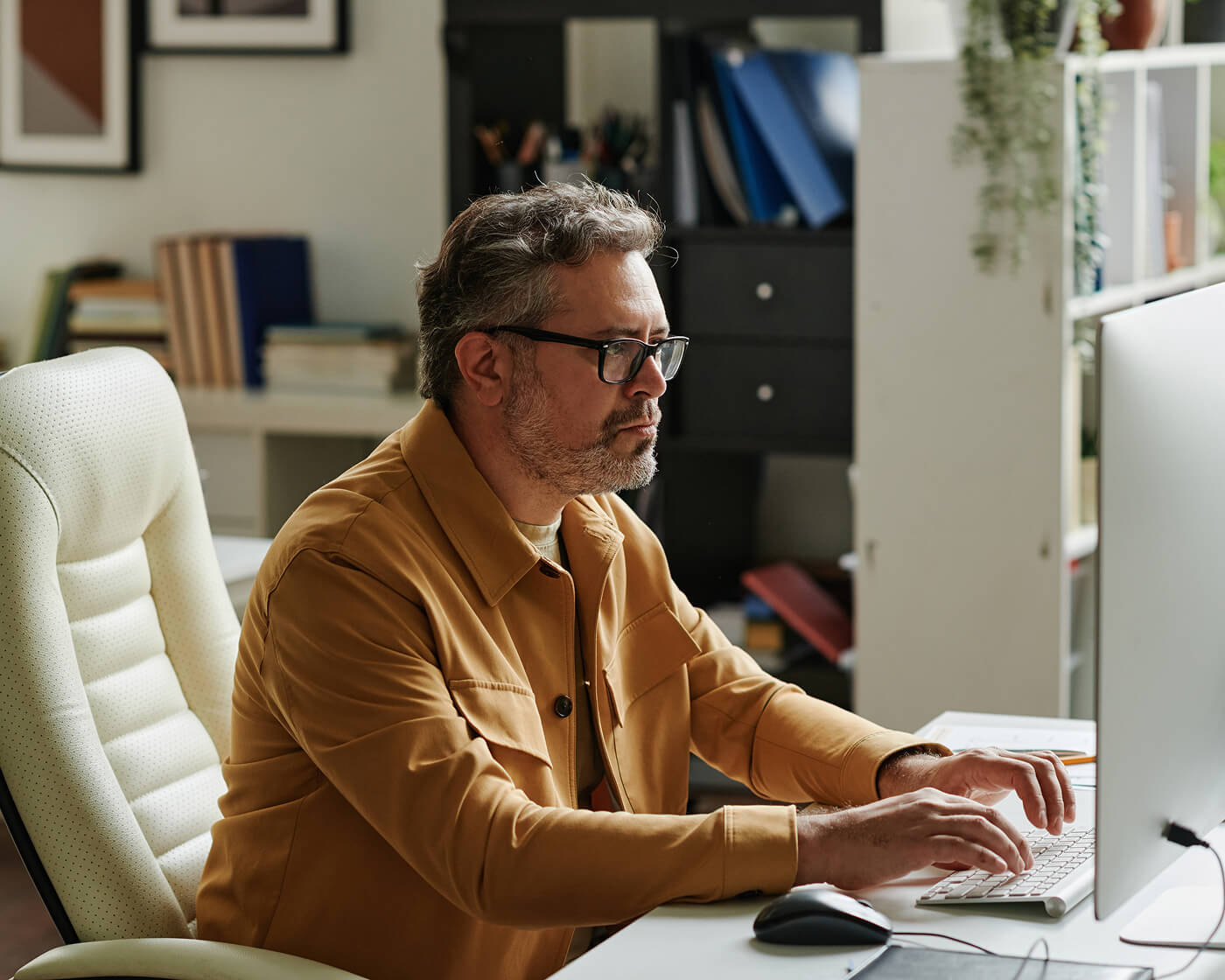 A man wearing glasses and a mustard jacket typing on a desktop computer in a office