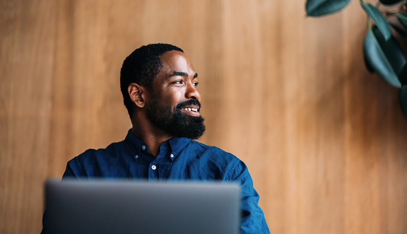 A smiling man using a laptop by a window.