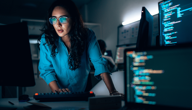 A woman wearing glasses typing on a keyboard while monitoring code on multiple computer screens