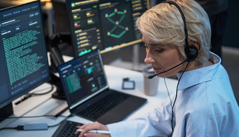 Woman wearing a headset monitoring security data on multiple screens.