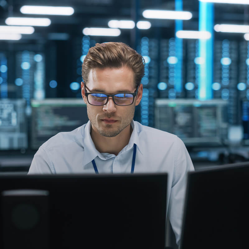 Man wearing glasses reviewing code on monitors in a server room.