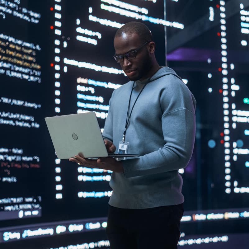 man-wearing-glasses-and-id-badge-holding-laptop-in-front-of-glowing-code-displays