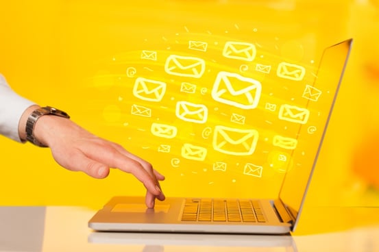 Image description: A woman's hand with her middle finger on the touchpad of an open laptop against a yellow background with numerous email symbols flowing from the laptop screen.