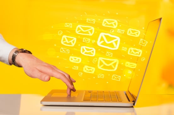 Image description: A woman's hand with her middle finger on the touchpad of an open laptop against a yellow background with numerous email symbols flowing from the laptop screen.