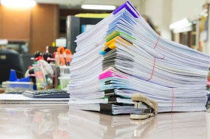 Image description: A stack of bound papers with multi-colored clips and binders sitting atop a desk with a miniature staple in the foreground. Behind the papers, there are numerous other office supplies, including a calculator, scissors, cups filled with pens and a tape dispenser. 