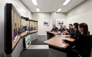 Image description: a rounded board table in a video conferencing room facing a screen depicting another team at a similar table that is conferencing in with the use of telepresence.