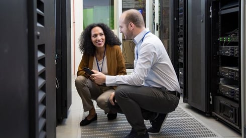 Image description: A woman and a man crouching in front of data center technology making direct eye contact and smiling.